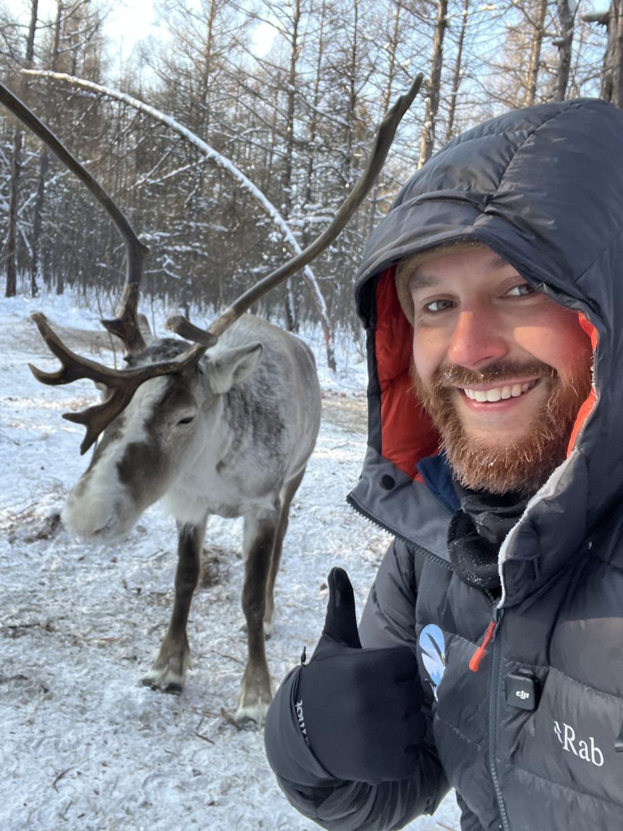John giving a thumbs up in a winter coat beside a reindeer
